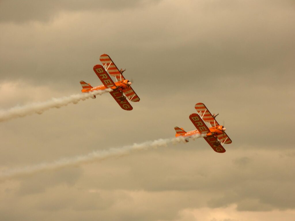 Two classic biplanes perform aerial stunts under cloudy skies, leaving smoke trails.